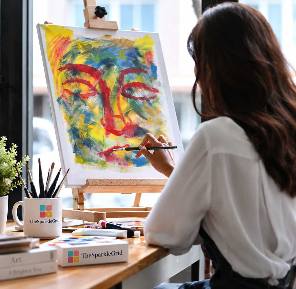 A woman painting a colorful abstract face on a canvas set on an easel, viewed from behind. She is wearing a white blouse and sitting at a wooden desk with art supplies, including a mug branded TheSparkleGrid.