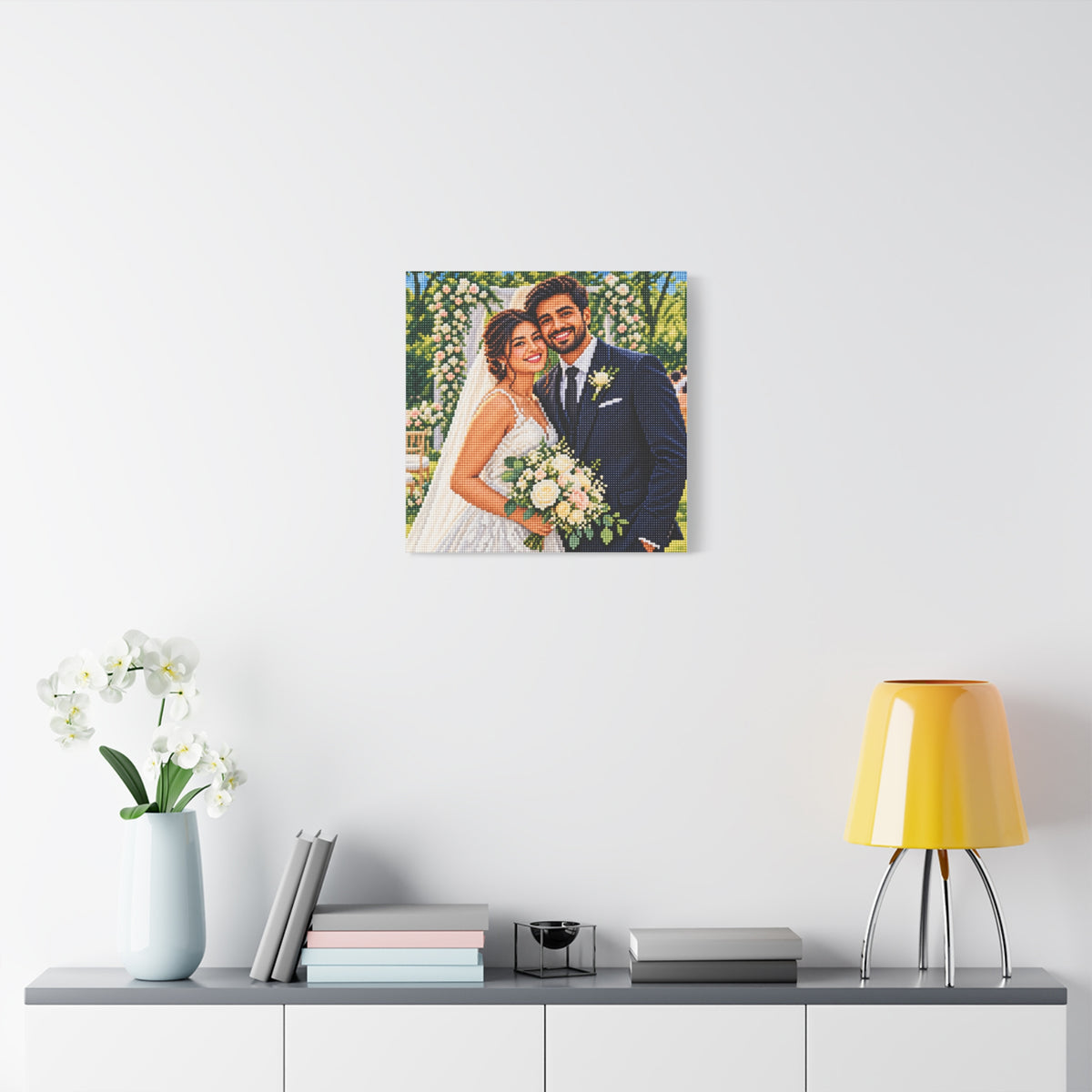 A square custom photo canvas print of a bride and groom, displayed on a white wall above a styled console table with a yellow lamp, books, and a vase of white flowers.